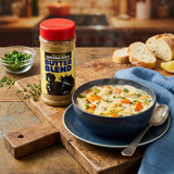 A bottle of Serendipity Butter Blend seasoning stands on a wooden cutting board beside a blue bowl of chicken and noodle soup with carrots, herbs, bread, and a spoon. The scene is set in a warm kitchen environment.