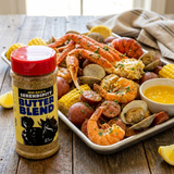 A bottle of Jess Hally's Serendipity Butter Blend seasoning is shown in the foreground next to a large seafood boil platter on a rustic wooden table. The platter contains shrimp, crab legs, clams, red potatoes, sausage, corn, and a small bowl of yellow butter sauce.
